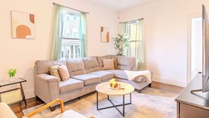 A living room with a sectional sofa, a round coffee table, a TV stand, and a ceiling fan. - Cozy 3Bed only 4min from tufts-10min from downtown Boston (Medford)