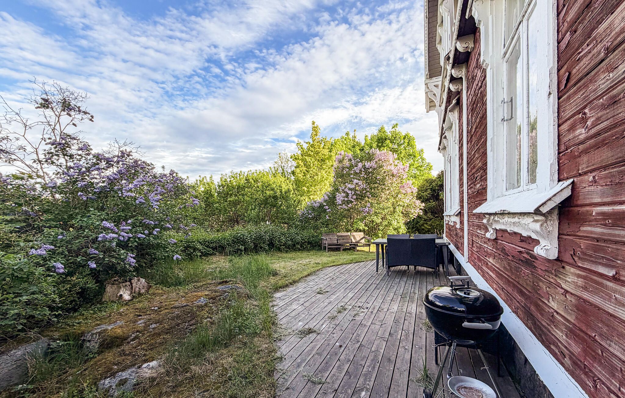 Une terrasse en bois équipée d’un barbecue et de mobilier d’extérieur, entourée d’une végétation luxuriante et d’arbustes en fleurs.
