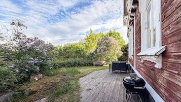 A wooden deck with a barbecue grill and outdoor furniture, surrounded by lush greenery and flowering bushes.