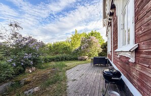Un terrazzo in legno con una griglia per barbecue e mobili da esterno, immerso nel verde e circondato da cespugli fioriti.