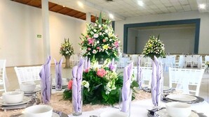 Una mesa de banquete dispuesta con platos blancos, cubiertos y servilletas moradas, rodeada de centros de mesa florales.