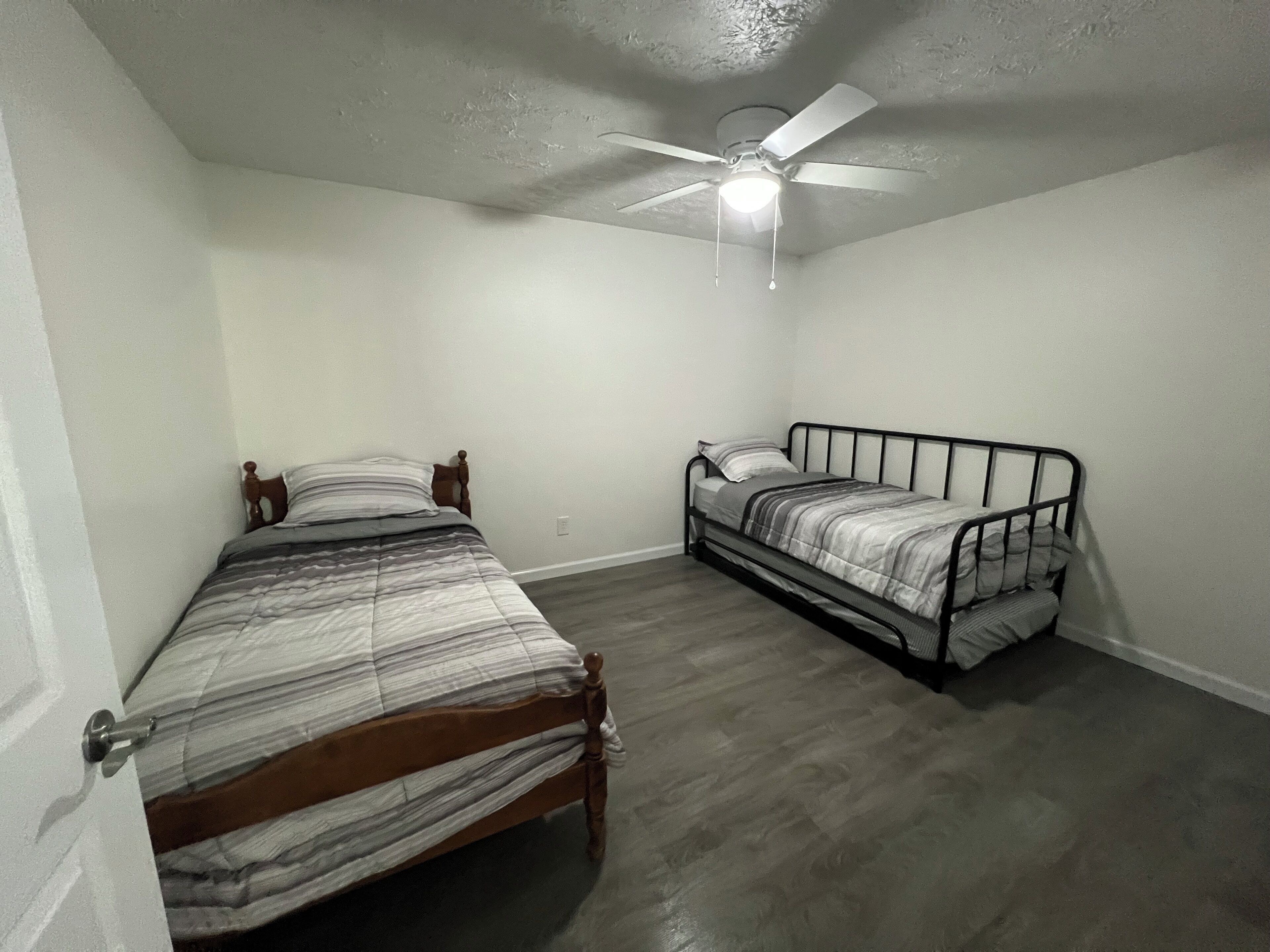 Un dormitorio con dos camas, una con estructura de madera y otra de metal, un ventilador de techo y una lámpara.