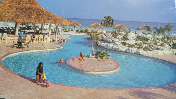 A pool area with lounge chairs and umbrellas, surrounded by palm trees and a view of the ocean. - Bahamas resort with 2 bedrooms, 2 bathrooms, full kitchen, living room, beach (Freeport)