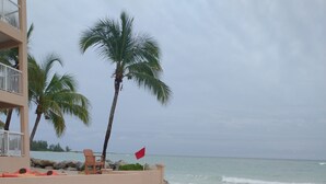 A beach with lounge chairs and a flag, palm trees, and a building in the background. - Bahamas resort with 2 bedrooms, 2 bathrooms, full kitchen, living room, beach (Freeport)