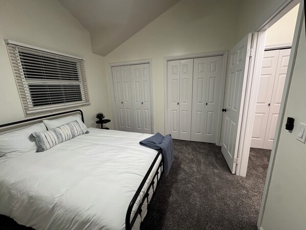 Une chambre à coucher avec un lit, une fenêtre avec des stores, une petite table avec une plante et une armoire à deux portes.