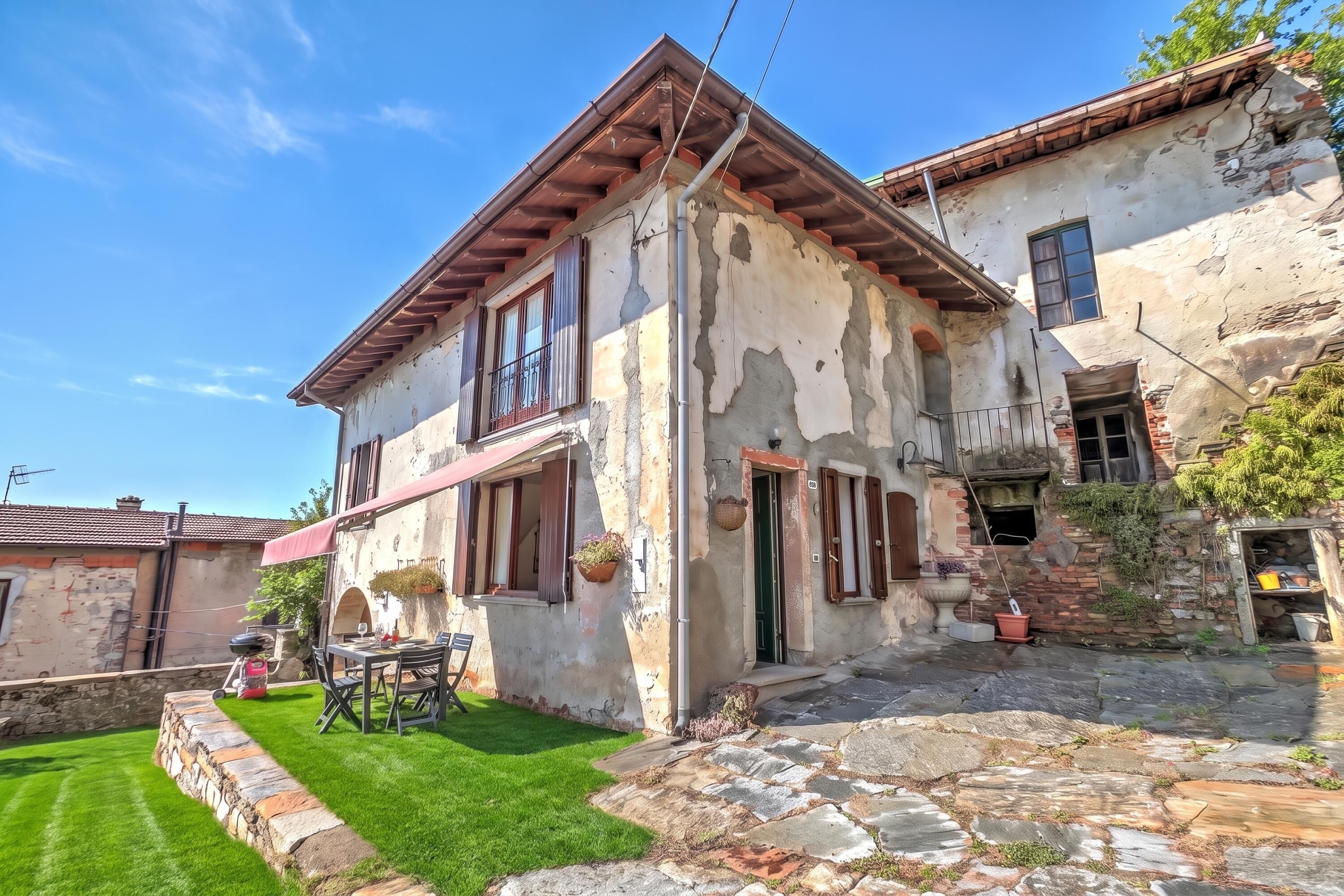 A two-story house with a stone facade, a balcony with a wooden railing, and a small garden area with a lawn and stone pathway.