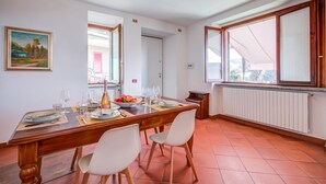 A dining area with a wooden table set for four, white chairs, a painting on the wall, and a window with a view. - Casa al Castello in Leggiuno With Garden (Leggiuno)