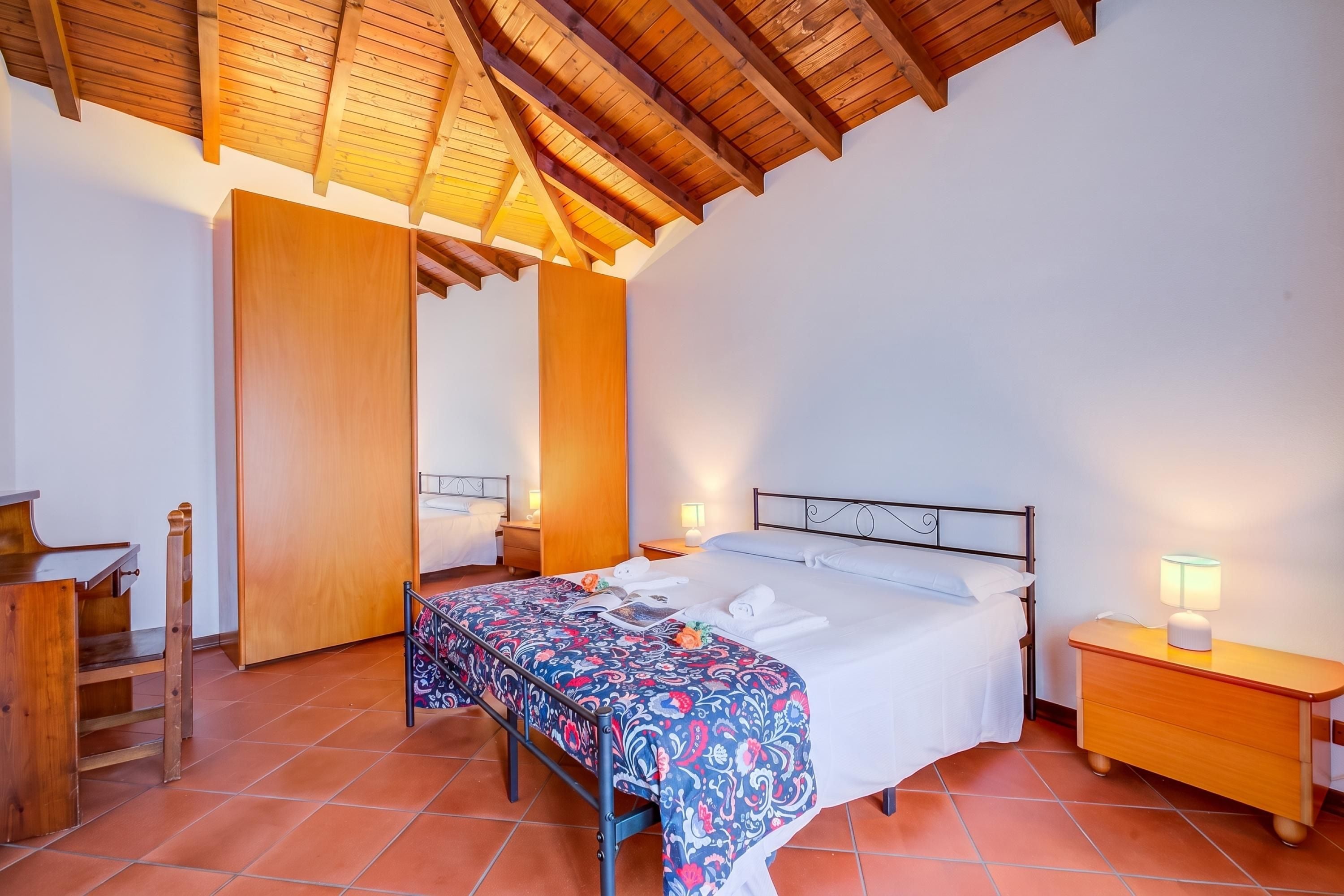 A bedroom with a wooden ceiling, a bed with a patterned bedspread, a nightstand with a lamp, and a wooden chair.