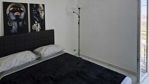 A modern bedroom with a black bedspread, a headboard with artwork, a floor lamp, and a large window. - Modern Hillside Retreat with Panoramic Views & Private Balcony with AC (Mussomeli)