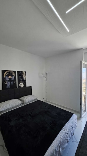 Ein modernes Schlafzimmer mit schwarzer Bettdecke, einem Kunstwerk-bezeichneten Kopfteil, einer Stehlampe und einem großen Fenster.