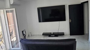 A modern living room with a flat-screen TV mounted on the wall, a dark sofa, and a large window providing natural light. - Modern Hillside Retreat with Panoramic Views & Private Balcony with AC (Mussomeli)