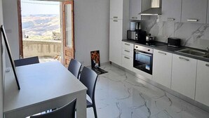 A modern kitchen with white cabinets, a built-in oven, and a microwave. There's a dining area with a table and chairs, and a view of the outdoors through a large window. - Modern Hillside Retreat with Panoramic Views & Private Balcony with AC (Mussomeli)