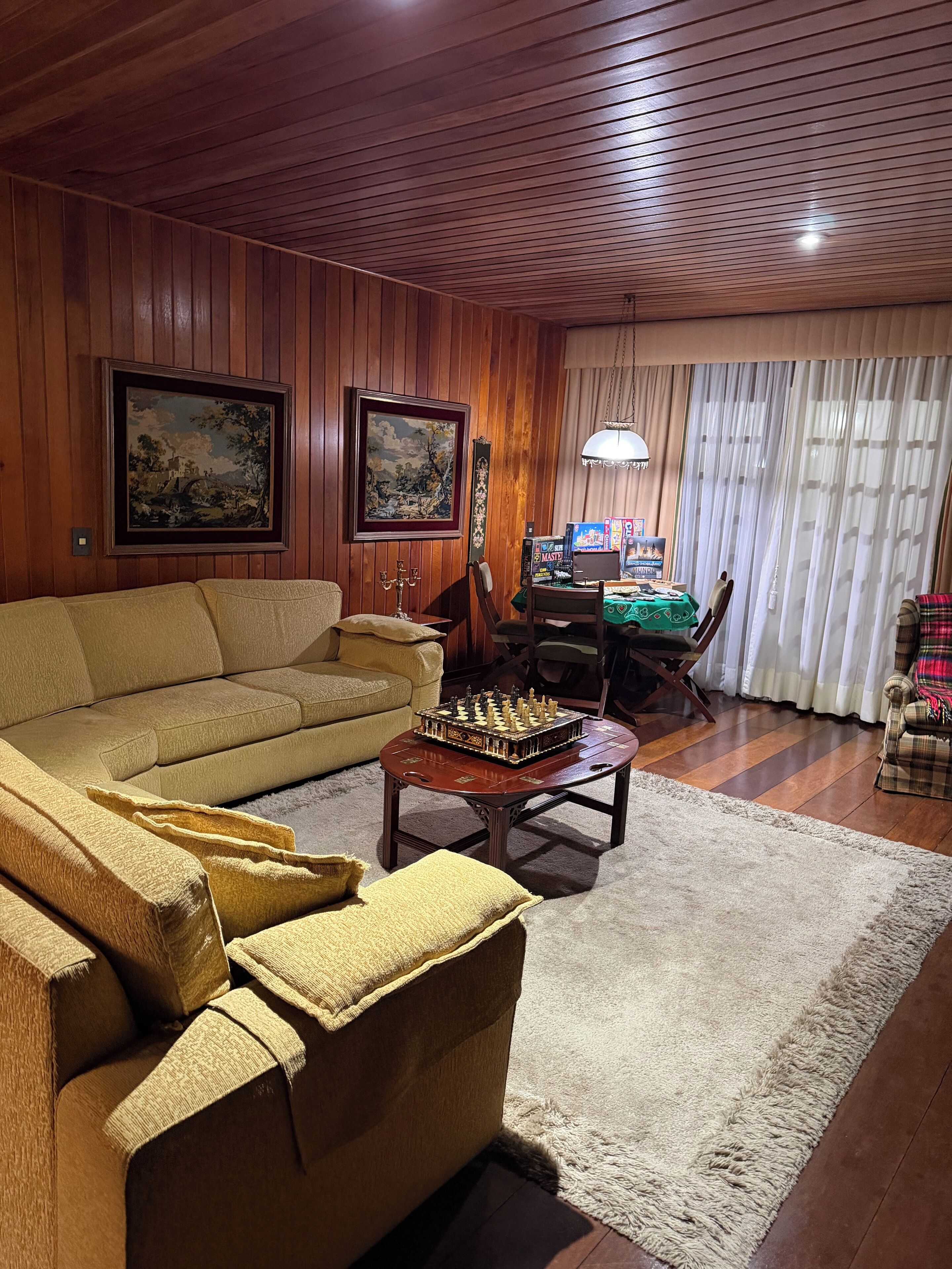 A cozy living room with wooden paneling, a sofa, a coffee table with a chess set, and two framed paintings on the wall.