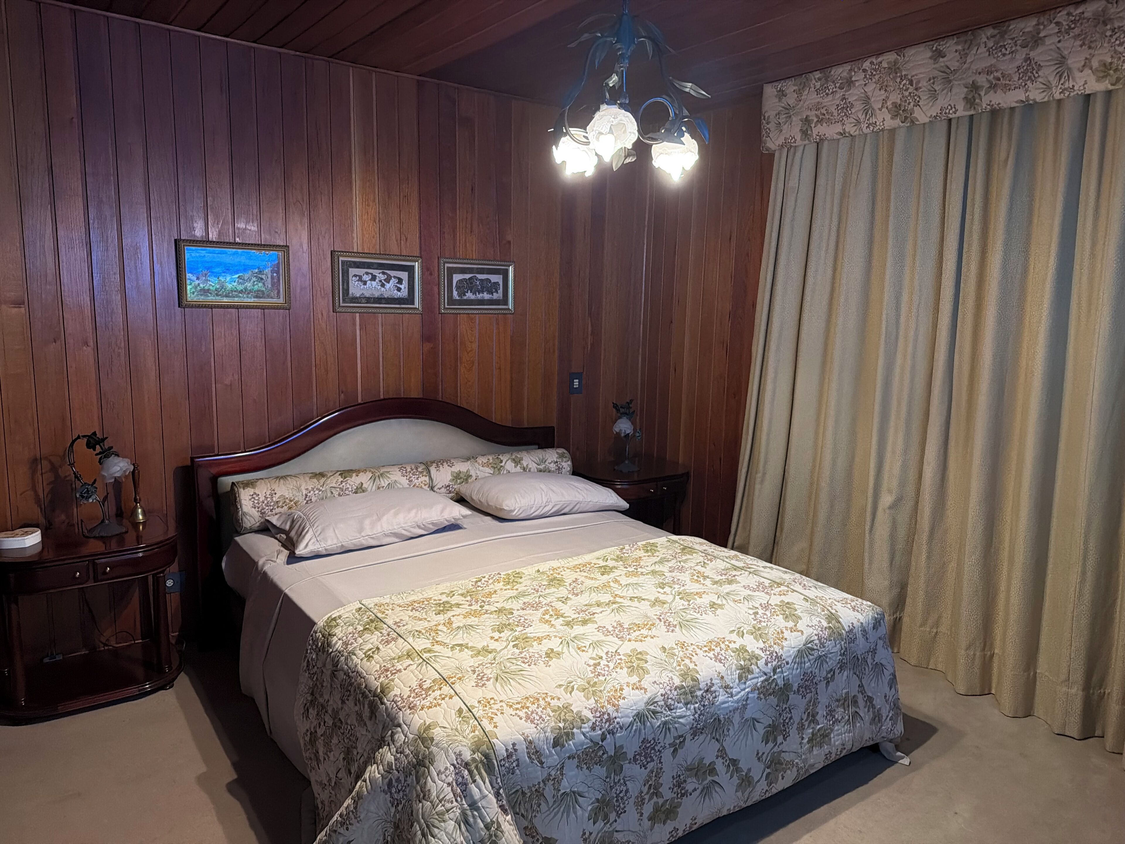 Une chambre avec un mur lambrissé en bois, un lit avec une literie à motifs floraux, une table de chevet avec une lampe et un lustre.