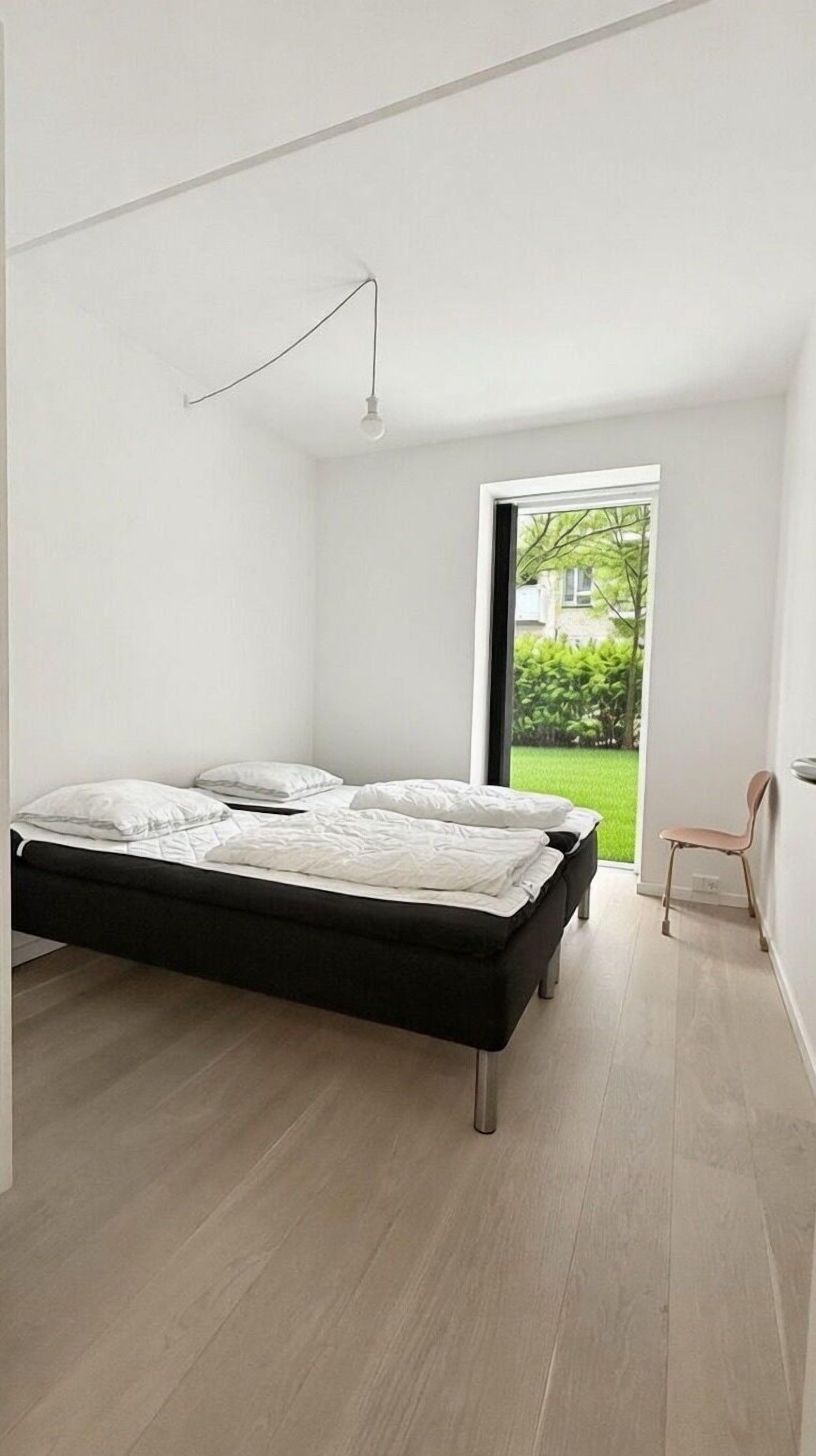 A modern bedroom with a large bed, a single hanging light, and a view of a green garden through a sliding door.