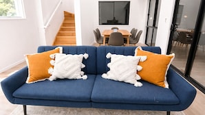 Un sofá azul con cojines blancos y naranja, una alfombra estampada y una escalera al fondo.
