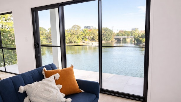 Una sala de estar moderna con un sofá azul y dos almohadones, con ventanas amplias que ofrecen vistas a un lago y follaje circundante.