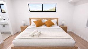 Un dormitorio moderno con una cama de madera, dos mesitas de noche, una ventana con vistas y un baño con lavamanos y espejo.