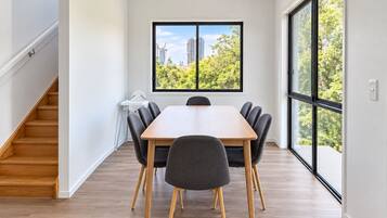 A modern dining area with a wooden table and chairs, a high window with a view of buildings and trees, and a staircase on the left.