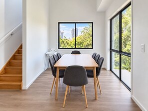 A modern dining area with a wooden table and chairs, a high window with a view of buildings and trees, and a staircase on the left. - Luxe Living in GC – Your Stylish Escape (Mermaid Waters)