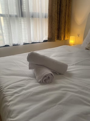 Ein ordentlich bezogenes Bett mit weißen Leinen und einem zusammengerollten Handtuch, eine Nachttischlampe und Vorhänge mit Blumenmuster.