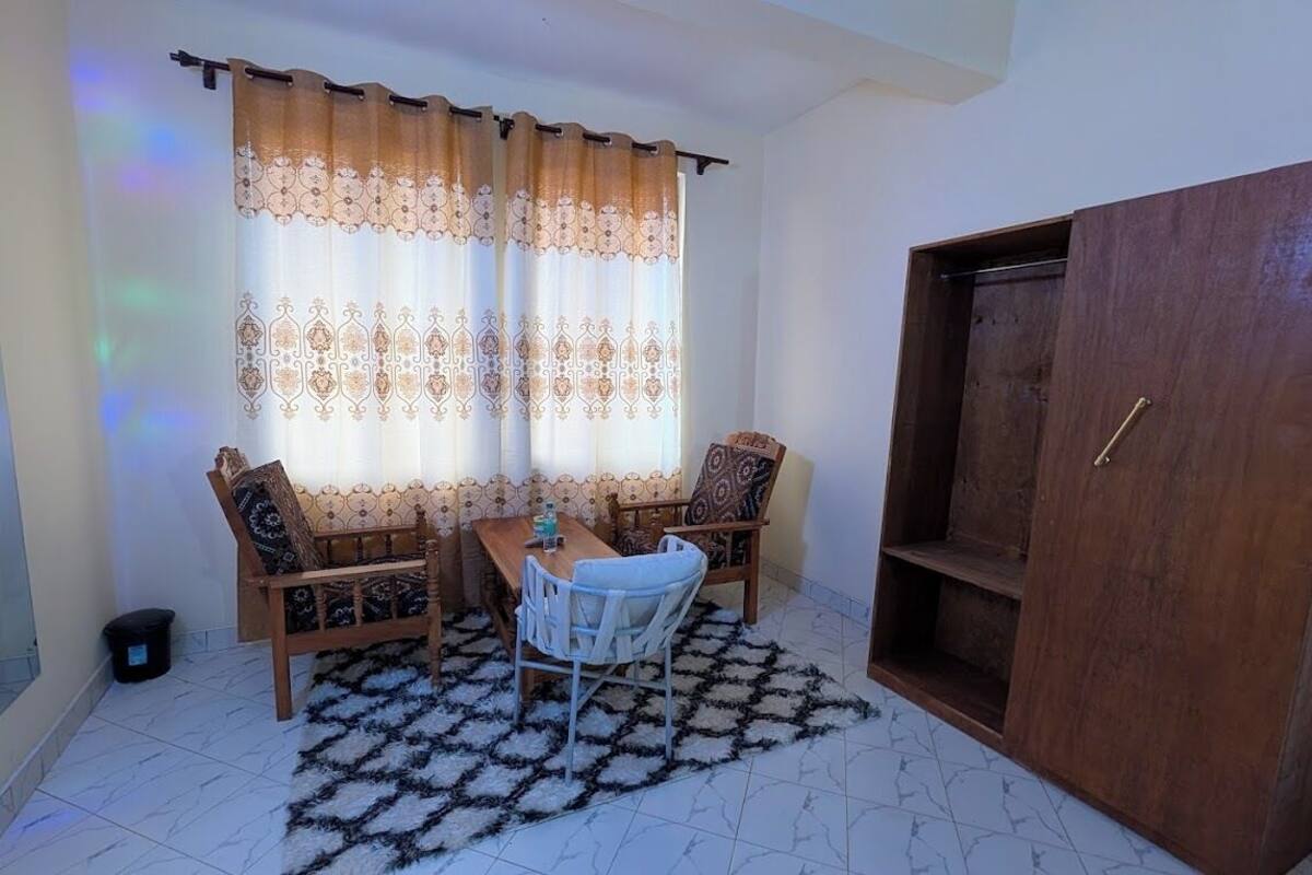 A room with a wooden wardrobe, a dining table with chairs, a patterned rug, and a window with curtains.