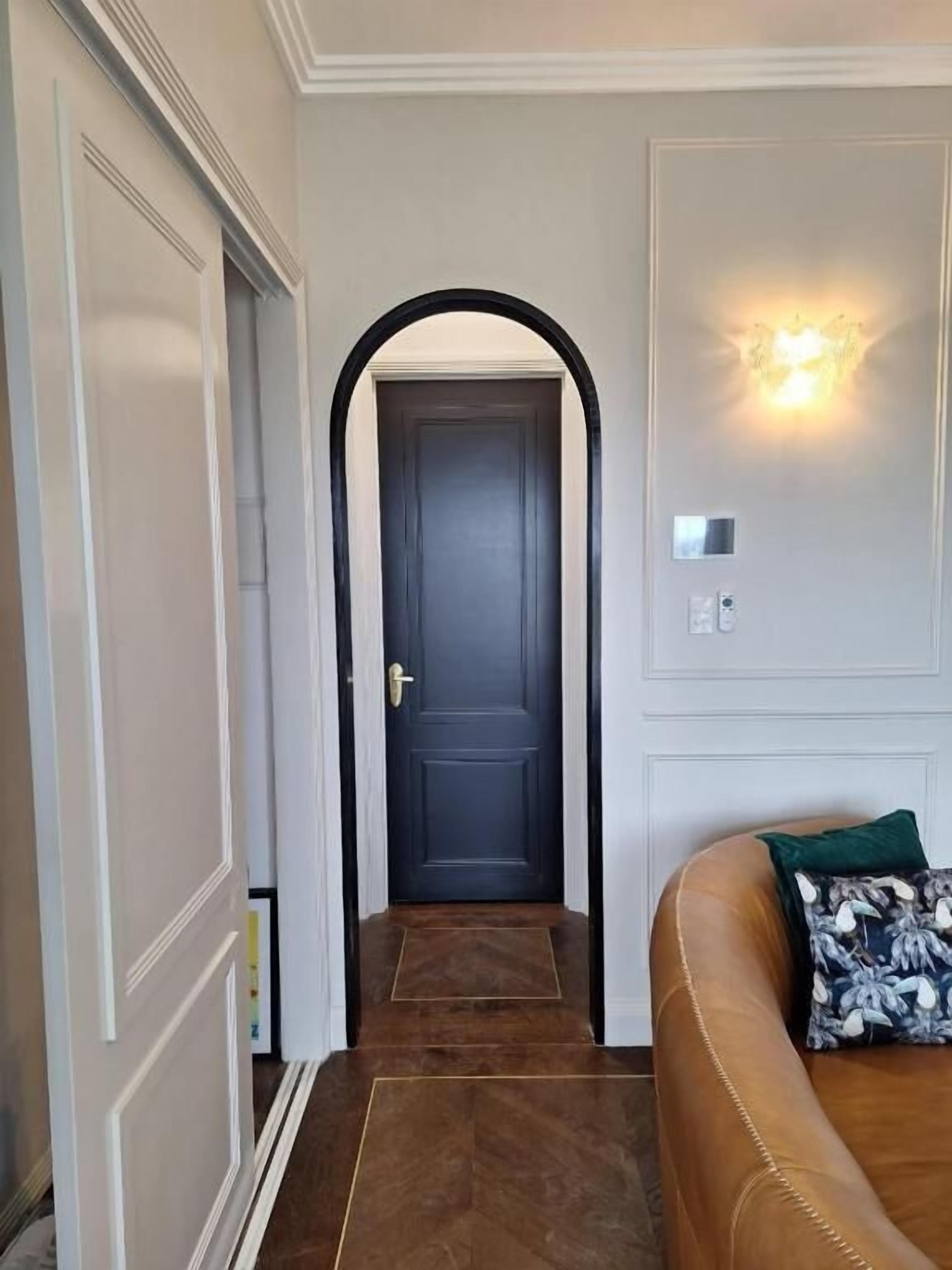 Un pasillo con una puerta azul oscuro, un sofá de cuero marrón y un cojín verde, y una lámpara de pared.