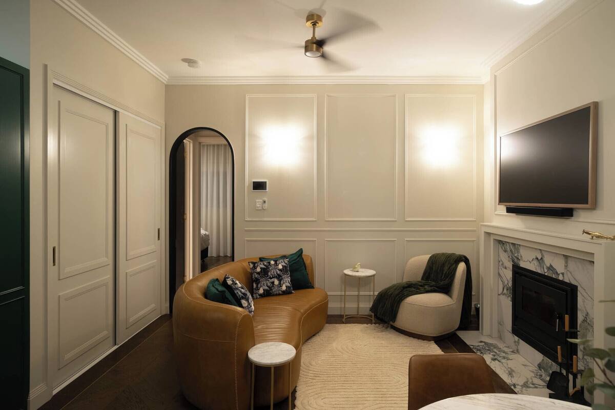 A living room with a brown leather sofa, a round coffee table, a white armchair, a fireplace, and a flat-screen TV mounted on the wall.