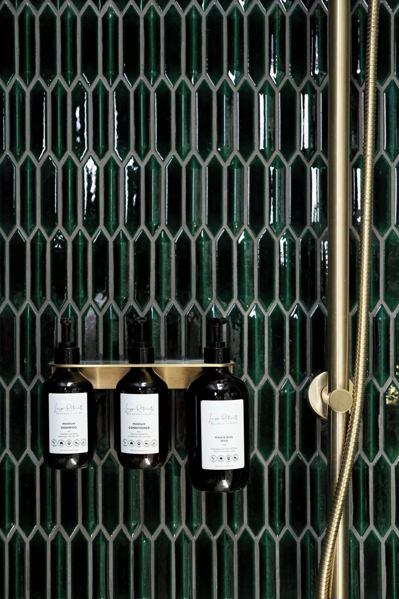 A bathroom with a green mosaic tile wall, a gold showerhead, and three bottles of Lush Retreat products.
