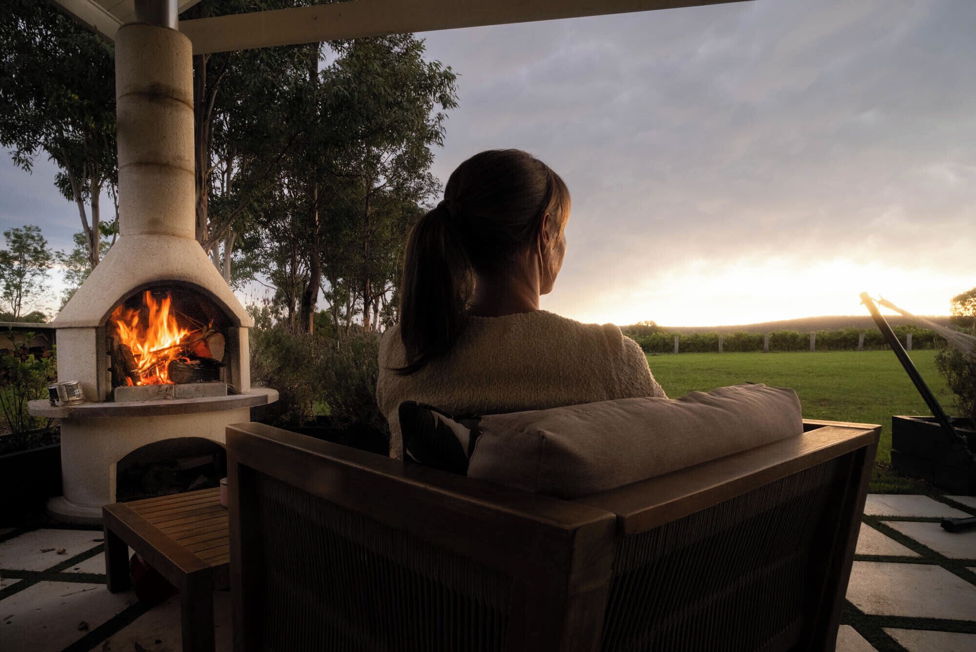 A person sitting on a chair facing a wood-burning stove with a fire burning inside, outdoors with a grassy field and trees in the background.