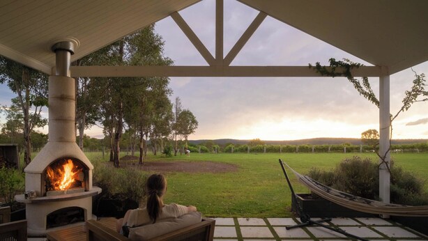 A covered outdoor seating area with a stone fireplace, a hammock, and a view of a vineyard. - Lusso Retreats (Moffatdale)