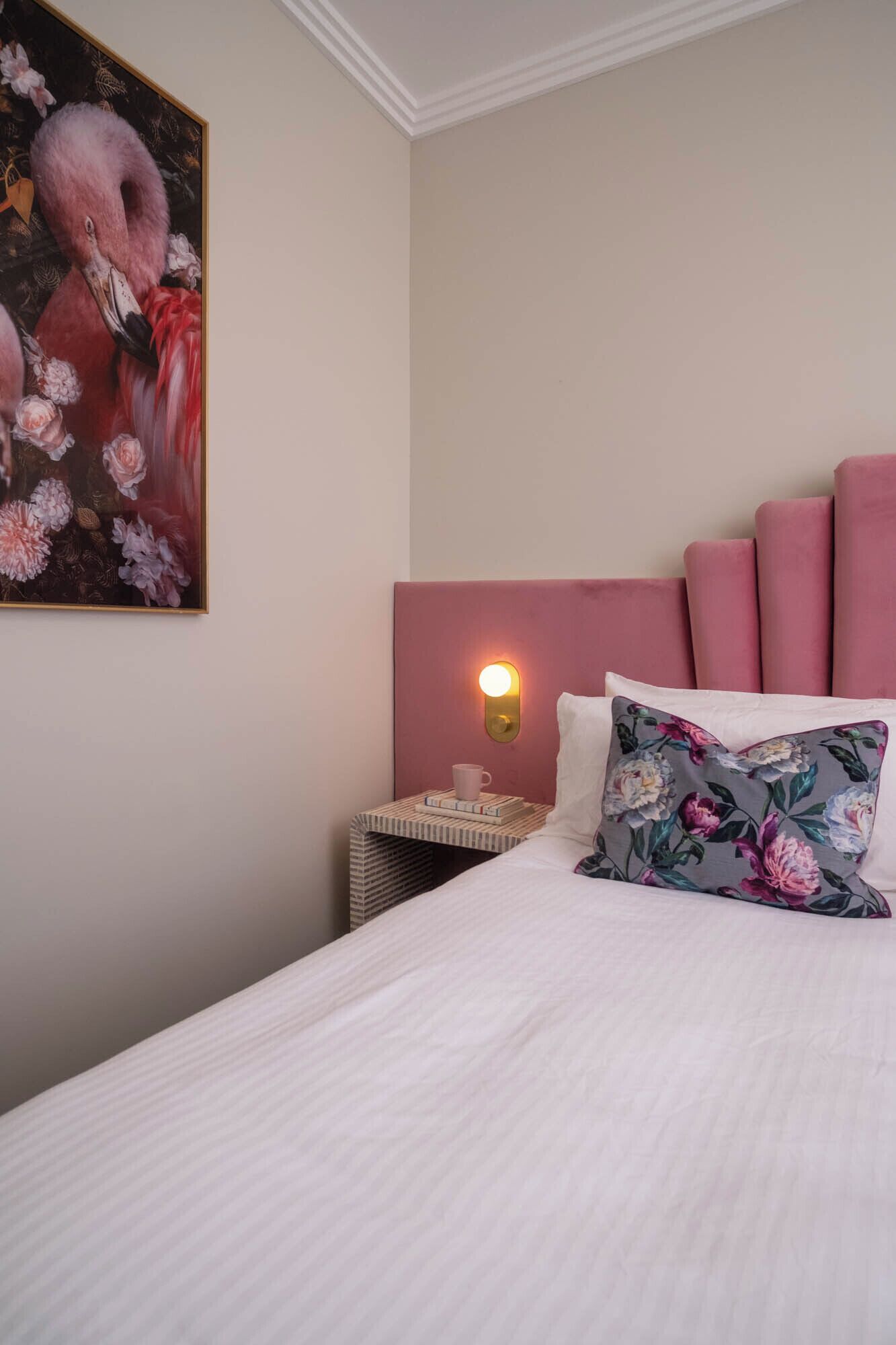 A neatly made bed with a floral pillow, a bedside table with a cup and books, and a wall-mounted light.