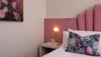 A neatly made bed with a floral pillow, a bedside table with a cup and books, and a wall-mounted light.