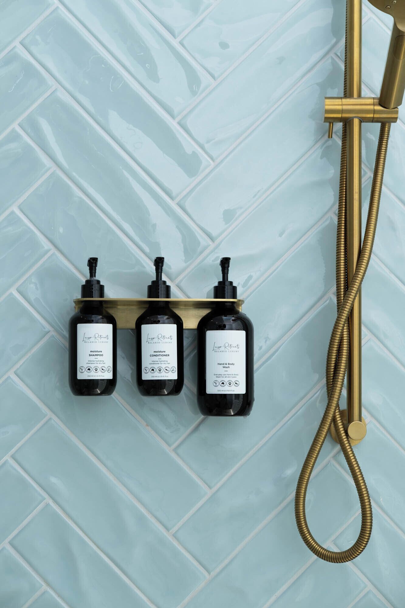 A bathroom with a blue tiled wall, a gold showerhead, and three bottles of shampoo, conditioner, and body wash on a shelf.