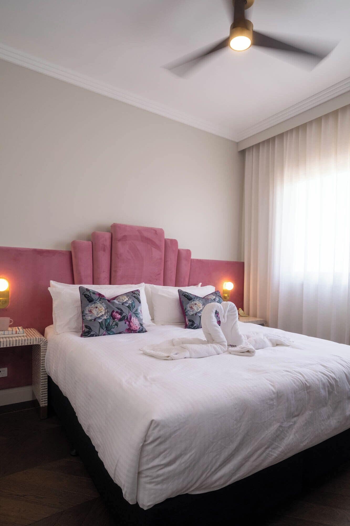 Una cama bien hecha con almohadas estampadas con motivos florales, un cabecero rosa y un ventilador de techo.