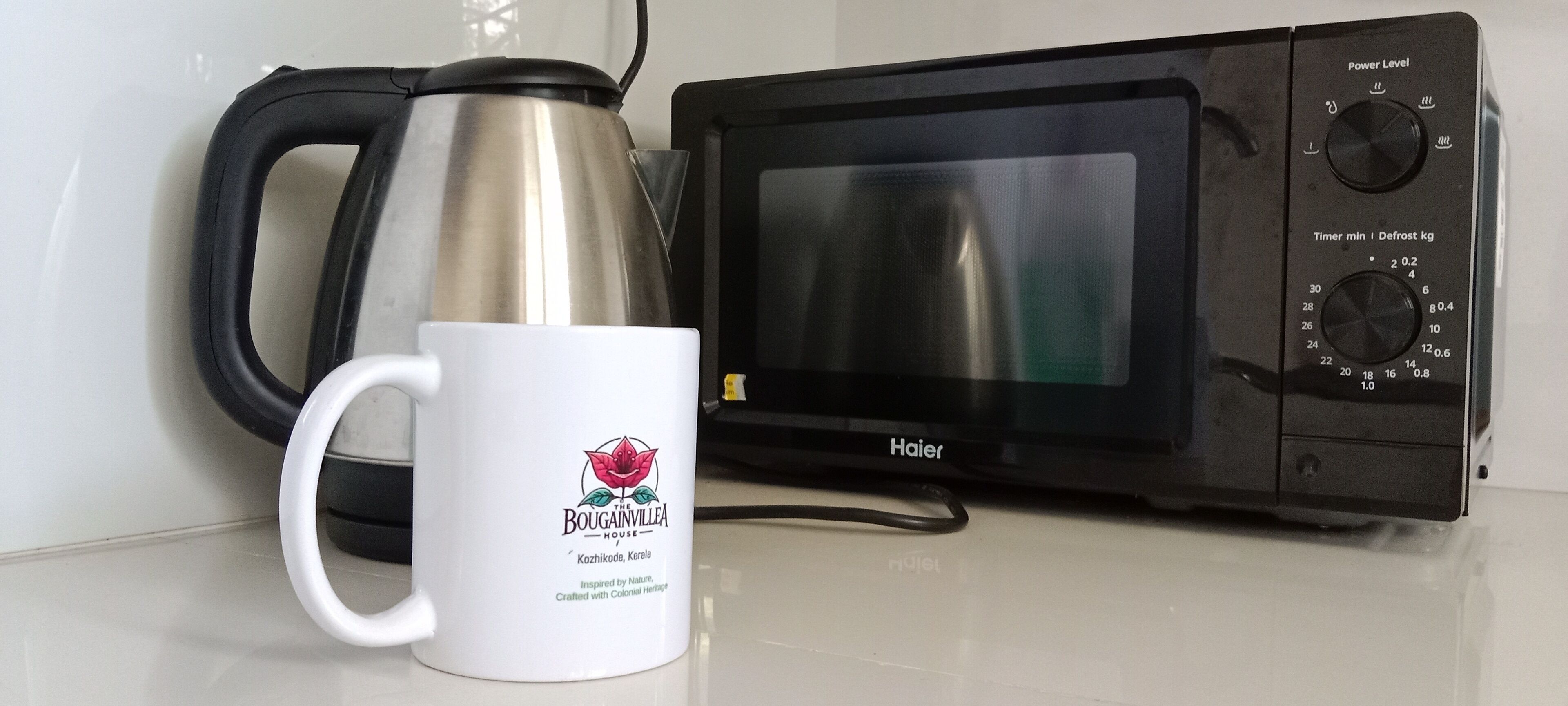A white mug with a logo and text, a stainless steel kettle, and a black Haier microwave on a white countertop.
