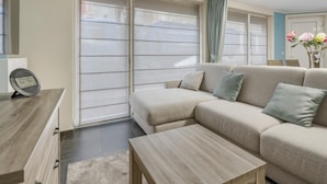 En moderne stue med en beige sofa, et træsofabord og et stort vindue med persienner.