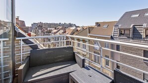 En balkong med möbler i rotting, ett metallräcke och utsikt över en stadssiluett med tak i terrakottafärg.