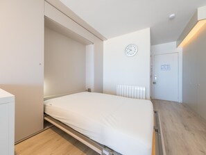 Une chambre à coucher moderne avec un lit, un radiateur blanc, une horloge au mur et une porte sur laquelle est affiché un avis.