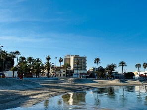 A beach with palm trees, a building with multiple floors, and a clear sky. - Hotel Albohera Playa by Belvilla Resorts-suite (San Javier)