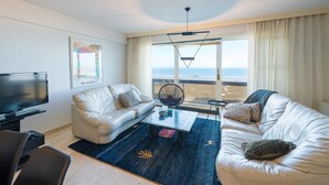 A modern living room with a large window, two white leather sofas, a coffee table, a TV, and a framed artwork. - Beautiful apartment on the 4th floor with garage (Middelkerke)
