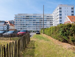 A multi-story residential building with balconies, a parking area with cars, and a wooden fence.