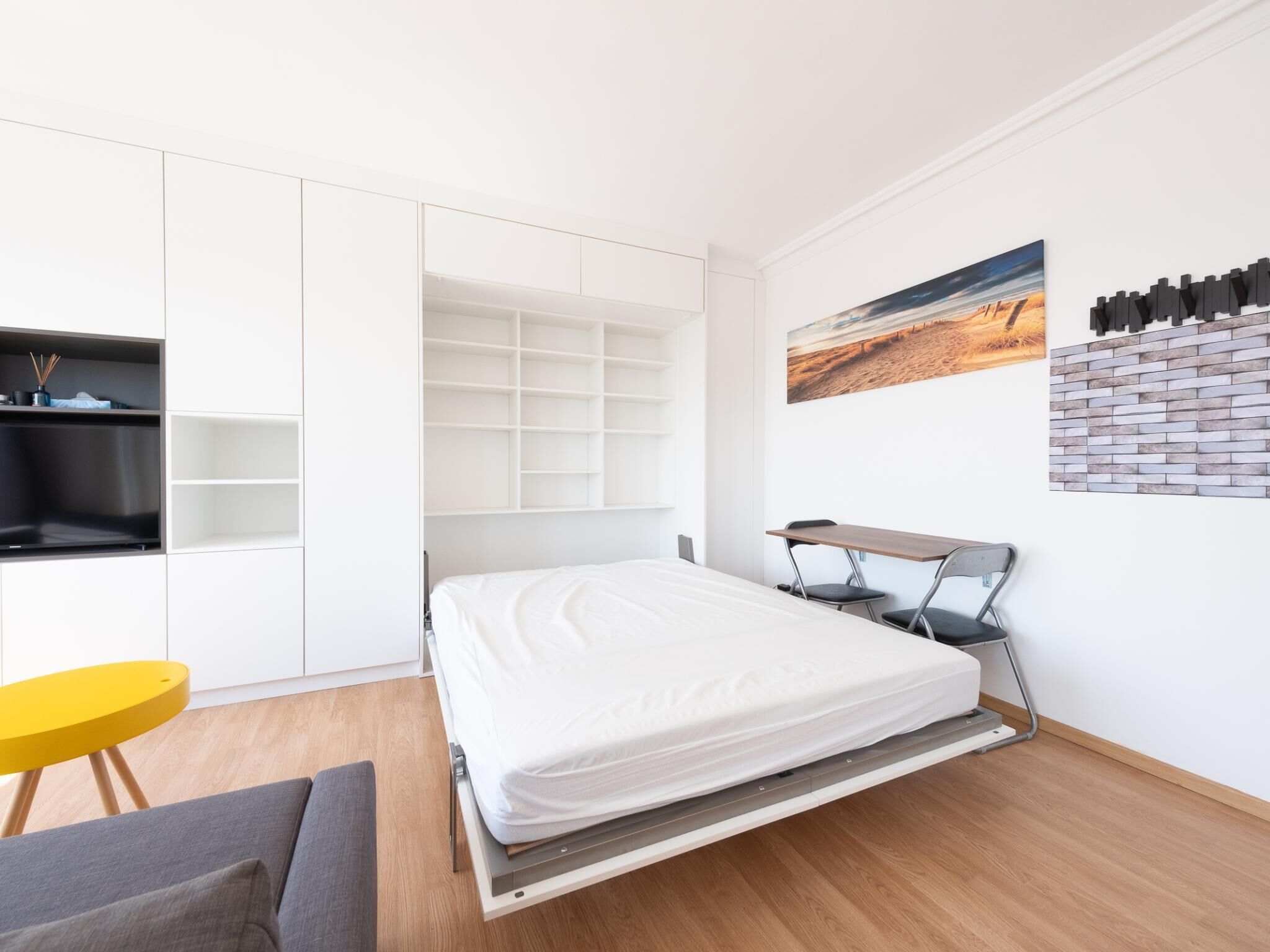 A modern bedroom with a bed, a desk, and a chair. There is a yellow stool and a gray sofa.