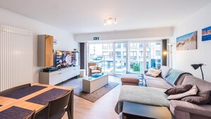 Een modern appartement met een eetgedeelte, een woonkamer met een bank en een televisie, een keuken met een kok, en grote ramen met uitzicht op de stad.