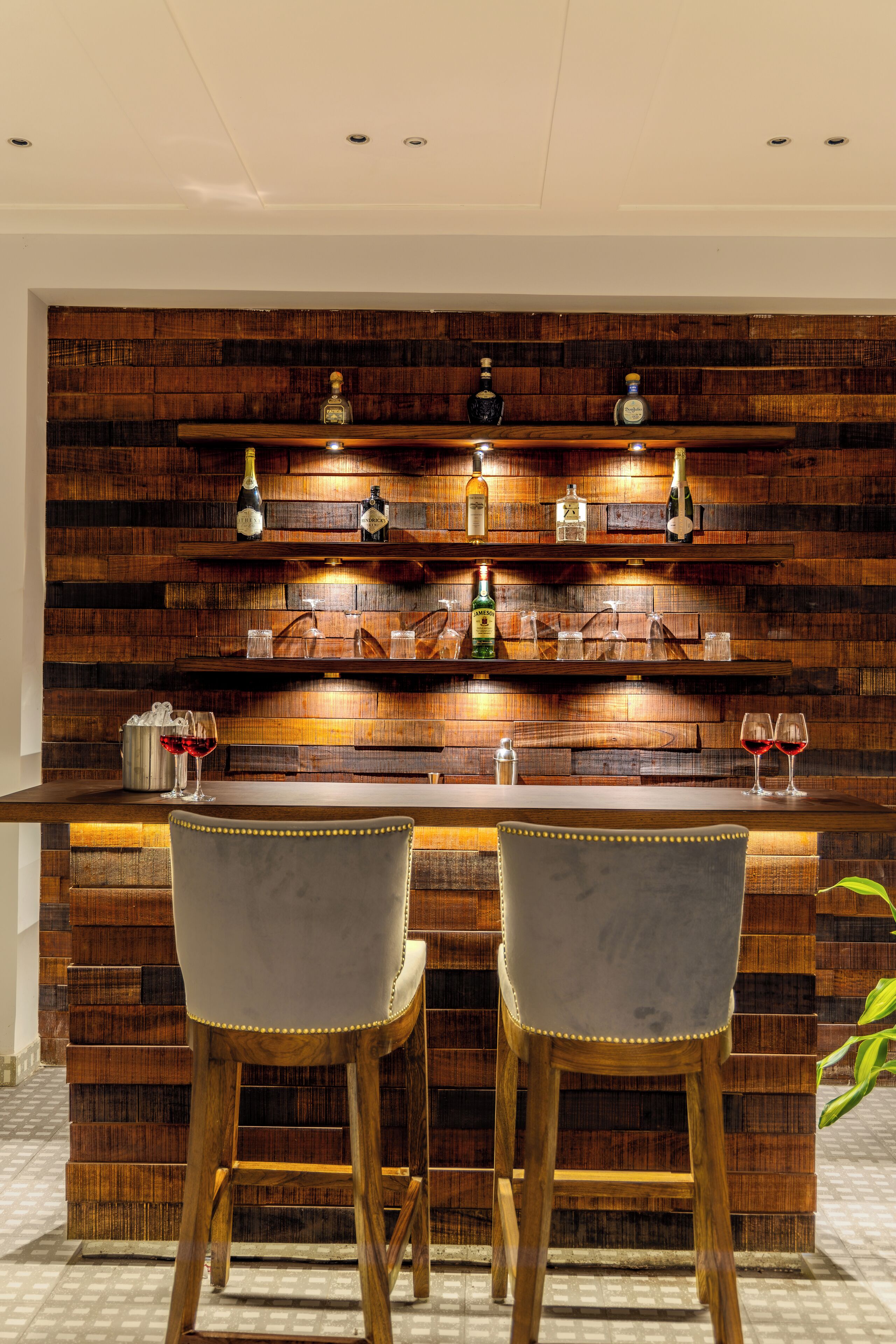 Zona de bar con paneles de madera, dos taburetes y estantes con botellas y vasos.