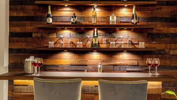 Zona de bar con paneles de madera, dos taburetes y estantes con botellas y vasos.