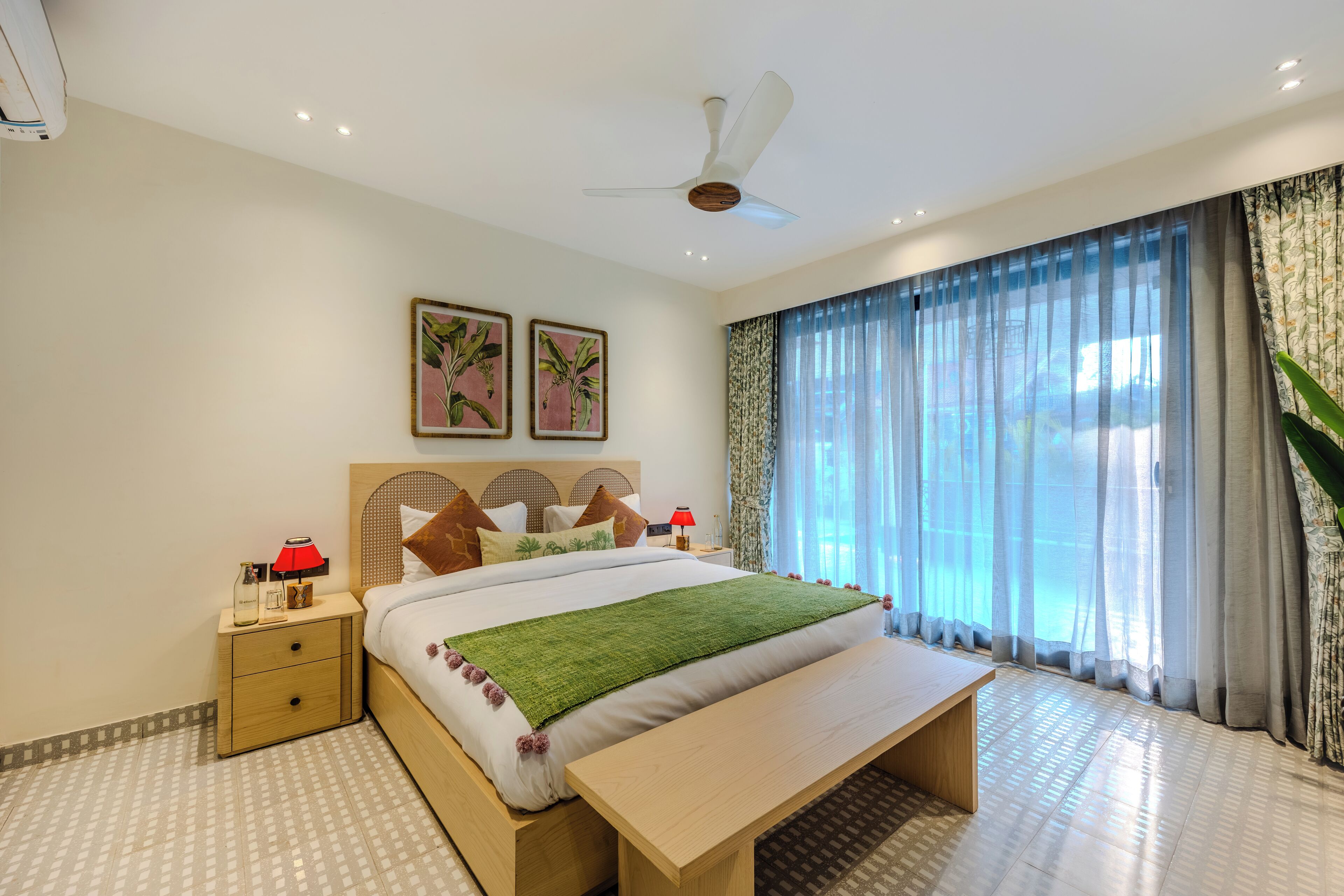 Quarto de hotel moderno com uma cama grande, mesa de cabeceira, ventilador de teto e uma janela ampla com cortinas.
