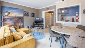 A living room with a large painting, a TV, a dining area with a table and chairs, and a kitchen visible through an open door. - Retro studio with sleeping area (Middelkerke)