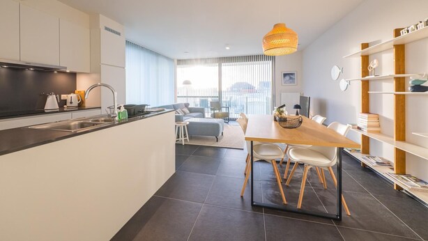 A modern kitchen with a central island, a dining area with a wooden table and chairs, and a living space with a sofa and a view of the city. - Spacious apartment with sunny terrace (Middelkerke)