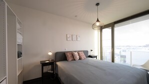 A modern bedroom with a large bed, a bedside table with a lamp, a wardrobe, and a city view through large windows. - Spacious apartment with sunny terrace (Middelkerke)